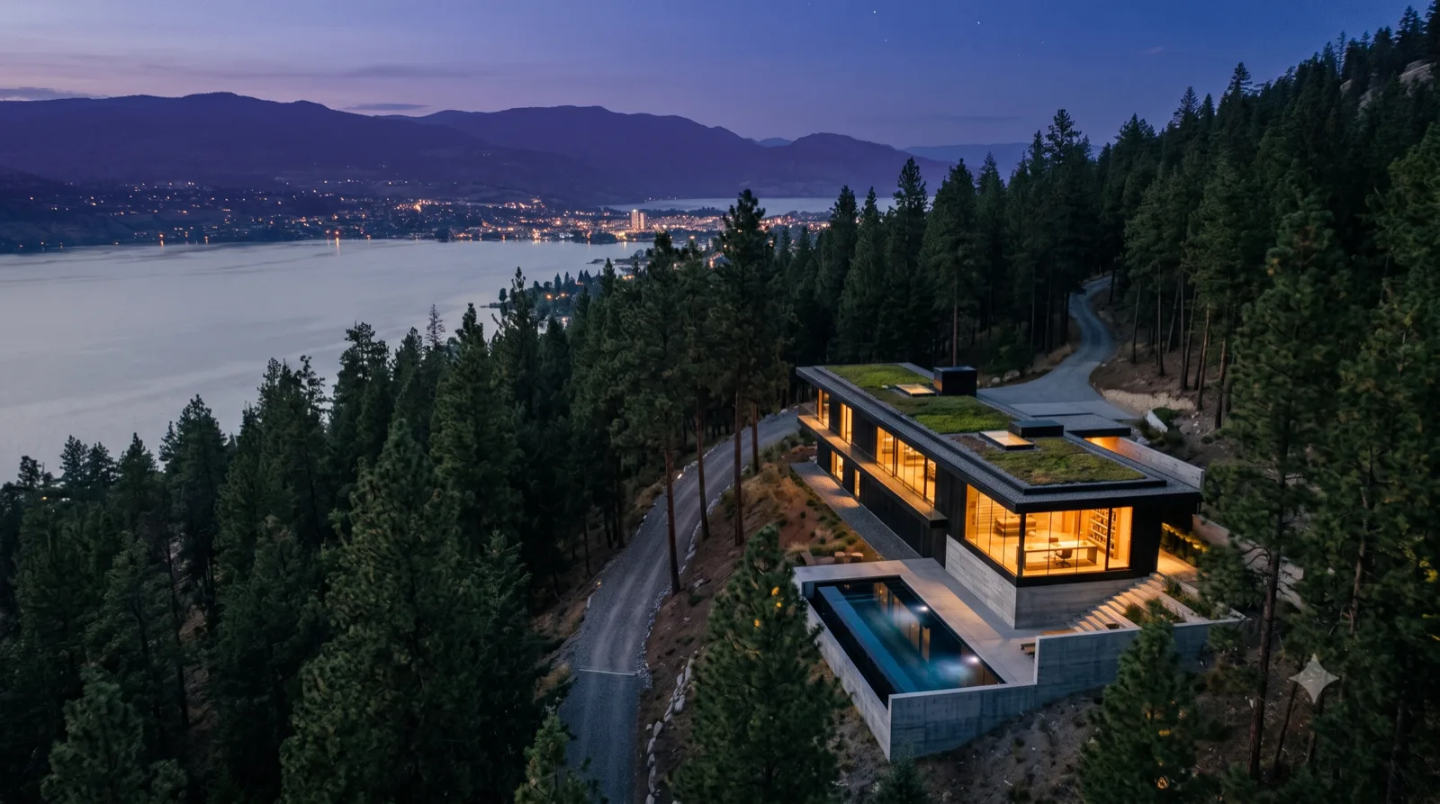 The Knox Mountain Perch — blue-hour aerial — green roofs among the treetops, infinity pool, Kelowna lights across the lake